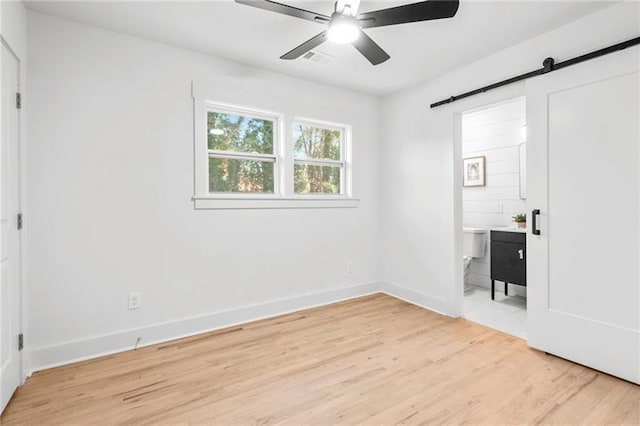 unfurnished bedroom with ceiling fan, a barn door, light wood-type flooring, and ensuite bath