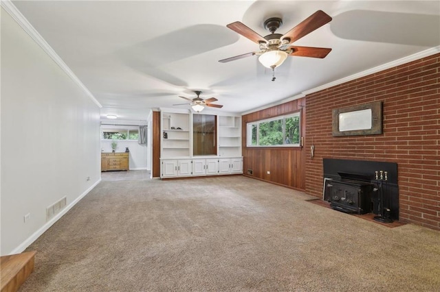 unfurnished living room with a wood stove, ornamental molding, carpet flooring, a ceiling fan, and baseboards