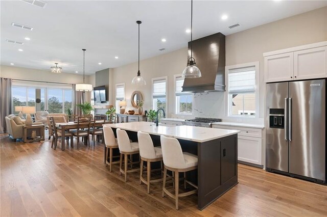 kitchen featuring appliances with stainless steel finishes, premium range hood, a wealth of natural light, a kitchen island with sink, and white cabinets