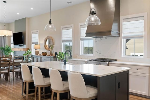 kitchen featuring hanging light fixtures, light hardwood / wood-style flooring, custom range hood, an island with sink, and white cabinetry