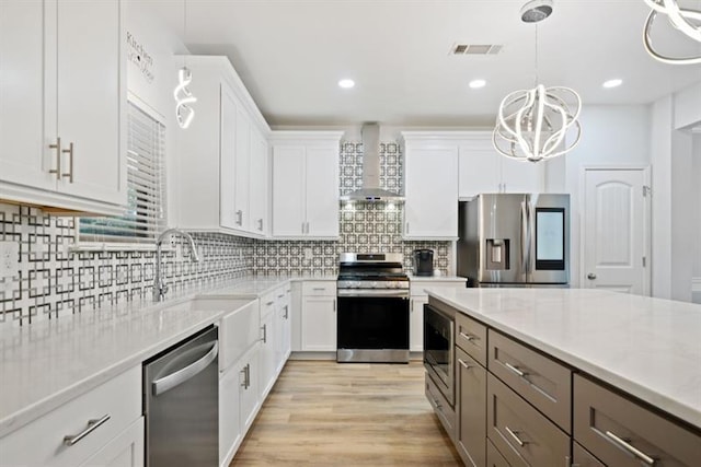 kitchen featuring appliances with stainless steel finishes, hanging light fixtures, wall chimney range hood, sink, and white cabinetry