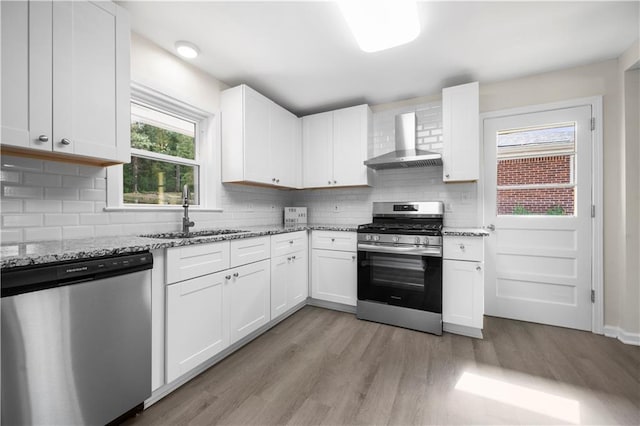 kitchen with sink, wall chimney range hood, light stone counters, white cabinets, and appliances with stainless steel finishes