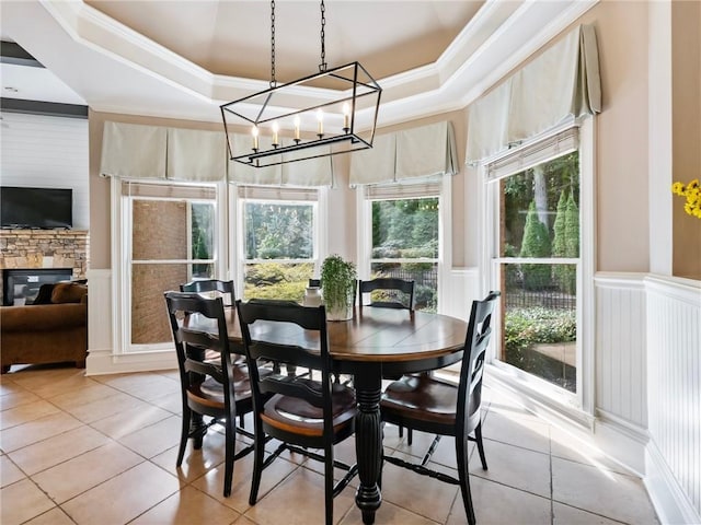 dining space with a stone fireplace, ornamental molding, light tile patterned floors, a raised ceiling, and an inviting chandelier