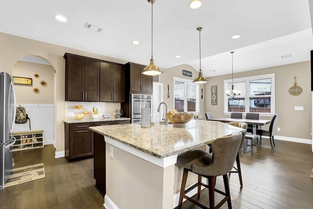 kitchen featuring dark hardwood / wood-style floors, a breakfast bar area, hanging light fixtures, a kitchen island with sink, and light stone countertops