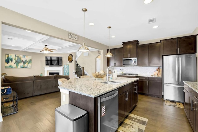 kitchen with a kitchen island with sink, stainless steel appliances, a sink, hanging light fixtures, and dark brown cabinets