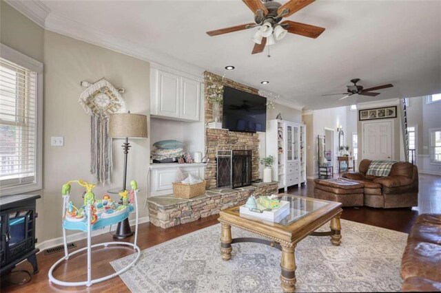 living area with dark wood finished floors, ornamental molding, a stone fireplace, and ceiling fan