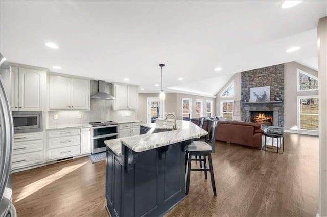 kitchen featuring hanging light fixtures, wall chimney exhaust hood, stainless steel appliances, an island with sink, and white cabinetry