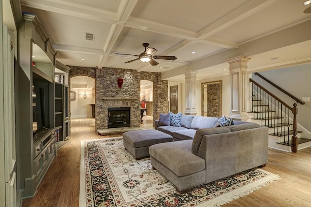 living room featuring wood finished floors, coffered ceiling, decorative columns, ceiling fan, and a stone fireplace