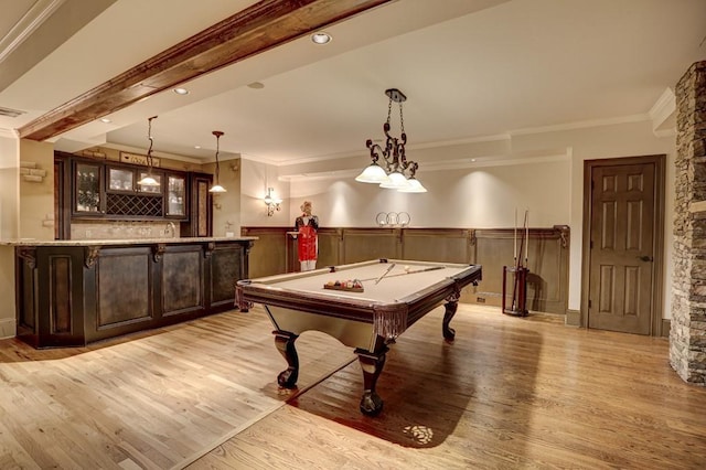 rec room with a wainscoted wall, beam ceiling, light wood-style flooring, bar area, and crown molding