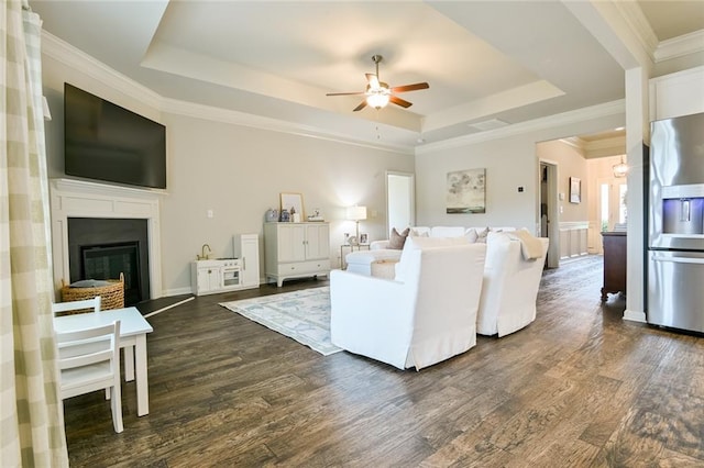 living room with a tray ceiling, ceiling fan, dark wood-type flooring, a fireplace, and ornamental molding