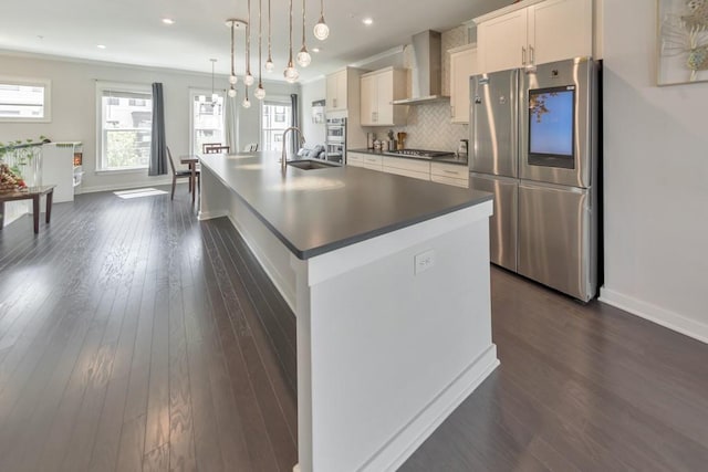 kitchen featuring wall chimney exhaust hood, a center island with sink, pendant lighting, stainless steel appliances, and white cabinets