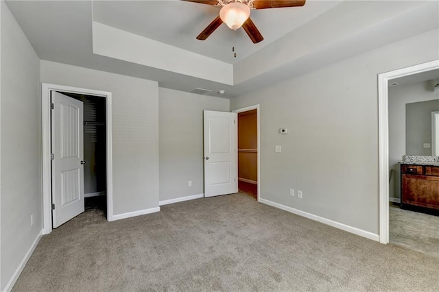 unfurnished bedroom with ceiling fan, a spacious closet, a tray ceiling, and light colored carpet