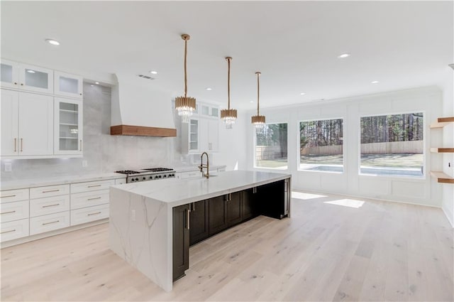 kitchen with custom exhaust hood, a kitchen island with sink, white cabinets, hanging light fixtures, and light wood-type flooring