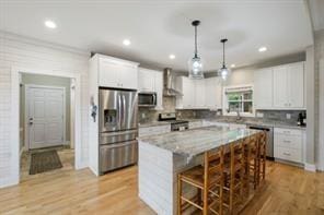 kitchen featuring wall chimney exhaust hood, decorative light fixtures, a kitchen island, white cabinetry, and stainless steel appliances