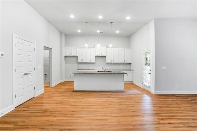 kitchen with white cabinetry, tasteful backsplash, hanging light fixtures, light wood-type flooring, and an island with sink