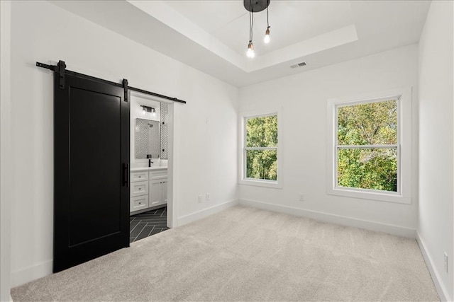 unfurnished bedroom with a tray ceiling, ensuite bathroom, a barn door, and carpet flooring