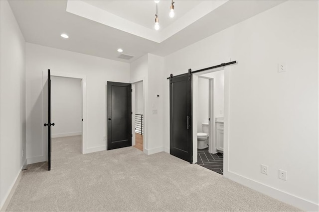 unfurnished bedroom featuring ensuite bathroom, a raised ceiling, light carpet, and a barn door