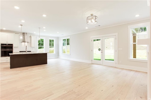 kitchen with multiple ovens, an island with sink, light wood-type flooring, french doors, and wall chimney exhaust hood