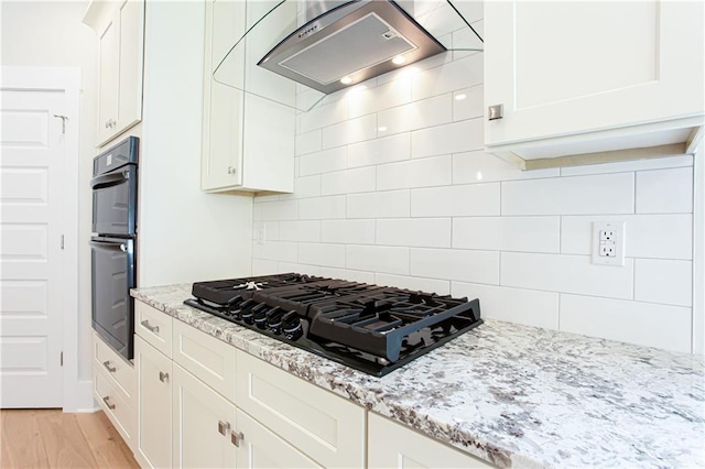 kitchen with light wood-type flooring, light stone counters, decorative backsplash, range hood, and black appliances