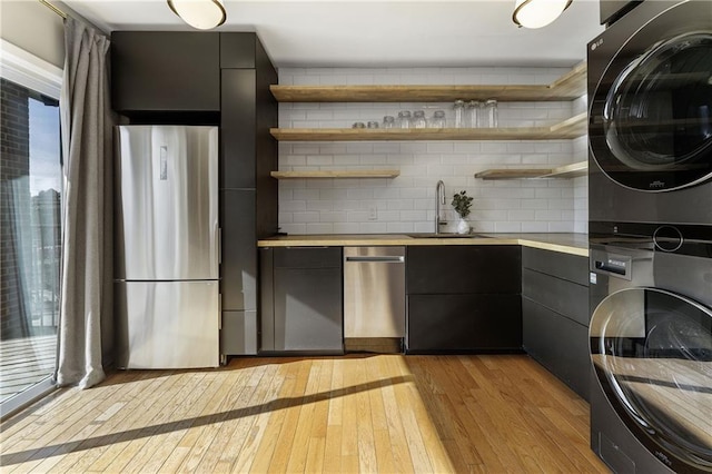 kitchen with sink, stainless steel appliances, stacked washing maching and dryer, light hardwood / wood-style floors, and backsplash