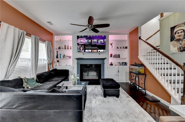 living area with dark wood-style floors, visible vents, stairway, a ceiling fan, and a glass covered fireplace