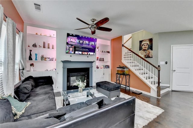 living area featuring visible vents, dark wood finished floors, a glass covered fireplace, ceiling fan, and stairs