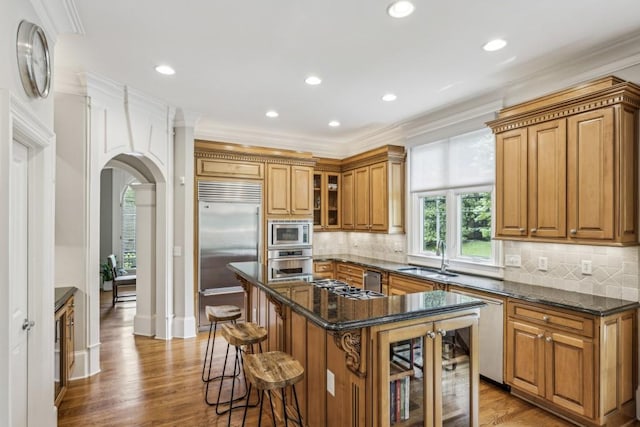 kitchen with built in appliances, a sink, brown cabinets, arched walkways, and a kitchen breakfast bar