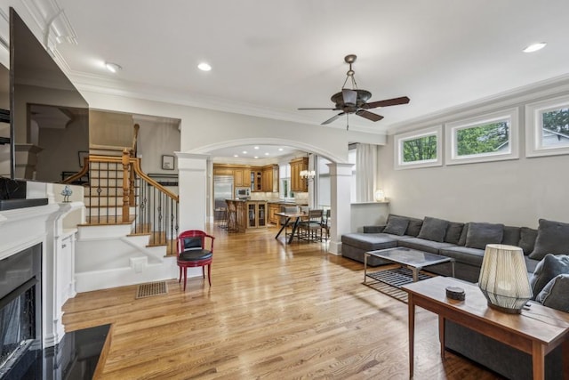 living room featuring a fireplace with flush hearth, ceiling fan, arched walkways, light wood-type flooring, and ornamental molding