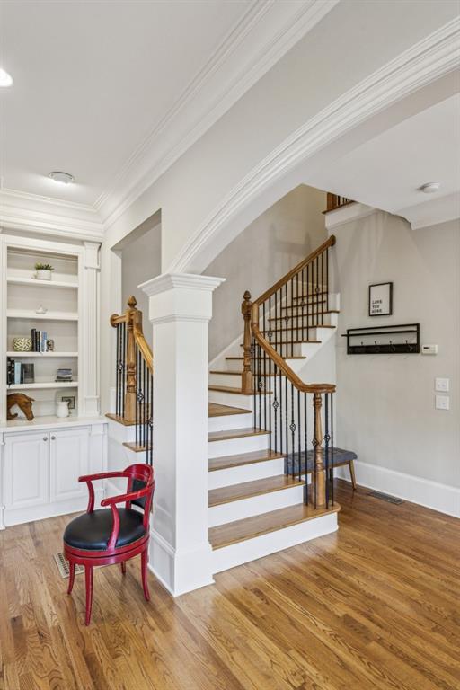 stairs featuring wood finished floors, crown molding, built in shelves, baseboards, and ornate columns