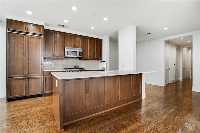 kitchen with decorative backsplash, stainless steel appliances, dark wood-type flooring, sink, and an island with sink