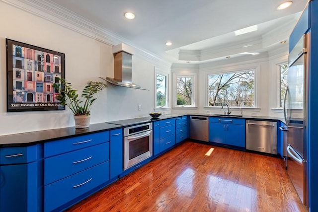 kitchen featuring blue cabinetry, stainless steel appliances, dark countertops, a sink, and wall chimney range hood