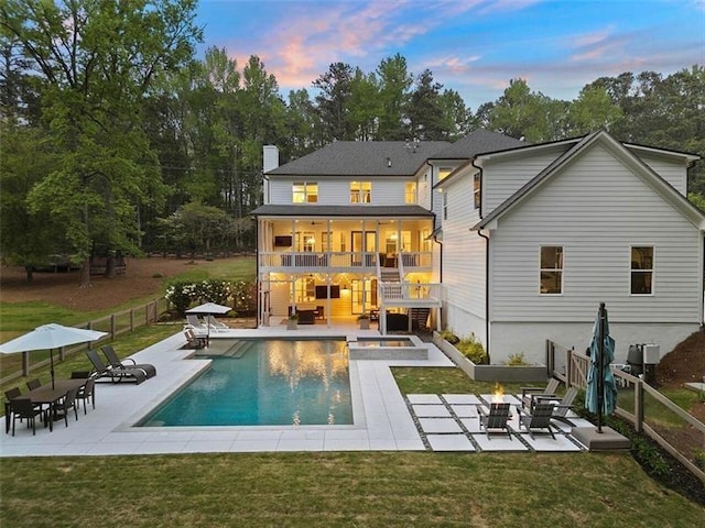 back house at dusk with a balcony, a swimming pool with hot tub, a patio area, and a fire pit