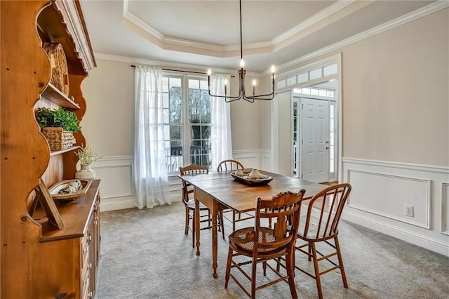 carpeted dining area with a wainscoted wall, a raised ceiling, an inviting chandelier, and crown molding