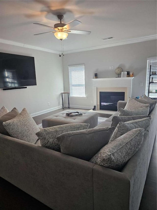 living area featuring baseboards, a glass covered fireplace, a ceiling fan, and crown molding