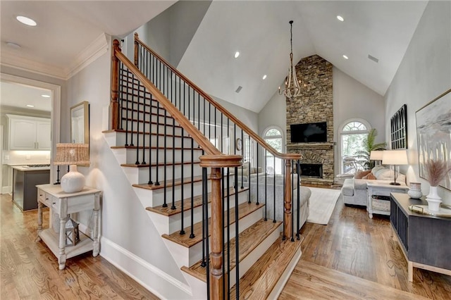 staircase featuring a chandelier, light hardwood / wood-style flooring, crown molding, a stone fireplace, and high vaulted ceiling