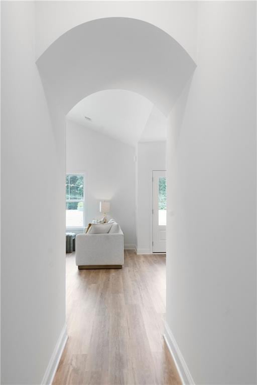 hallway with light wood finished floors, baseboards, arched walkways, and a wealth of natural light