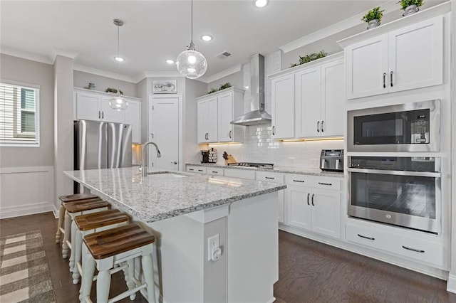 kitchen featuring white cabinets, wall chimney exhaust hood, a kitchen island with sink, stainless steel appliances, and crown molding