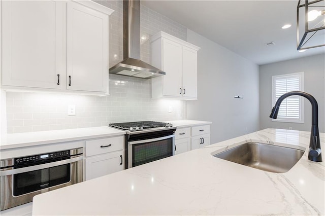 kitchen with wall chimney range hood, white cabinets, tasteful backsplash, a sink, and appliances with stainless steel finishes
