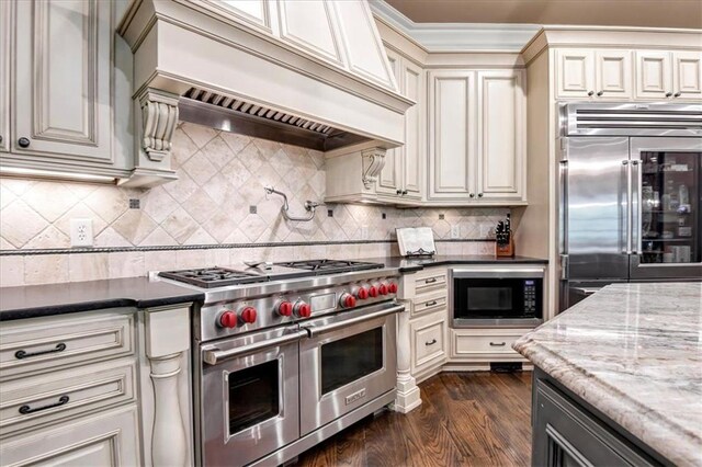 kitchen with backsplash, custom exhaust hood, dark hardwood / wood-style flooring, dark stone countertops, and built in appliances