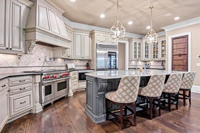 kitchen featuring dark stone counters, high quality appliances, a kitchen island with sink, dark hardwood / wood-style floors, and premium range hood