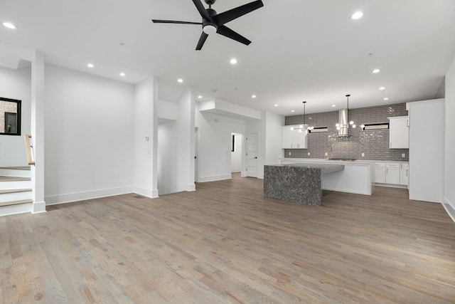 unfurnished living room featuring baseboards, stairway, recessed lighting, light wood-type flooring, and ceiling fan with notable chandelier