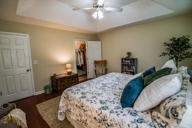 bedroom featuring a raised ceiling, dark wood finished floors, a walk in closet, baseboards, and a ceiling fan