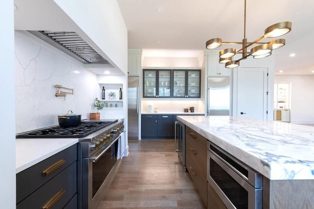 kitchen featuring built in appliances, wood finished floors, exhaust hood, backsplash, and glass insert cabinets