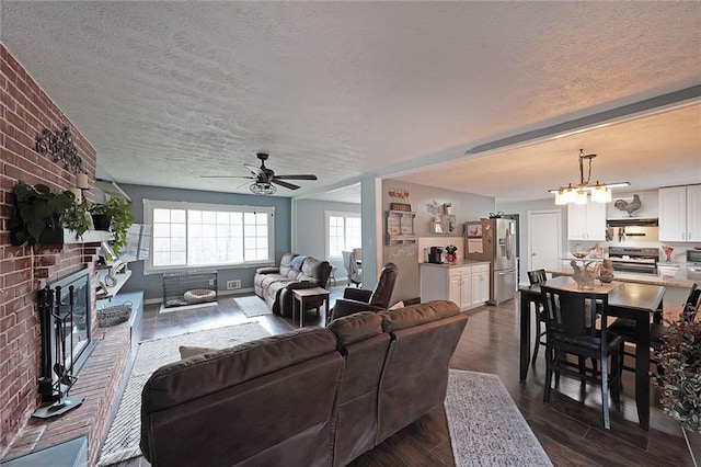 living room with brick wall, a brick fireplace, ceiling fan with notable chandelier, dark wood-style floors, and a textured ceiling