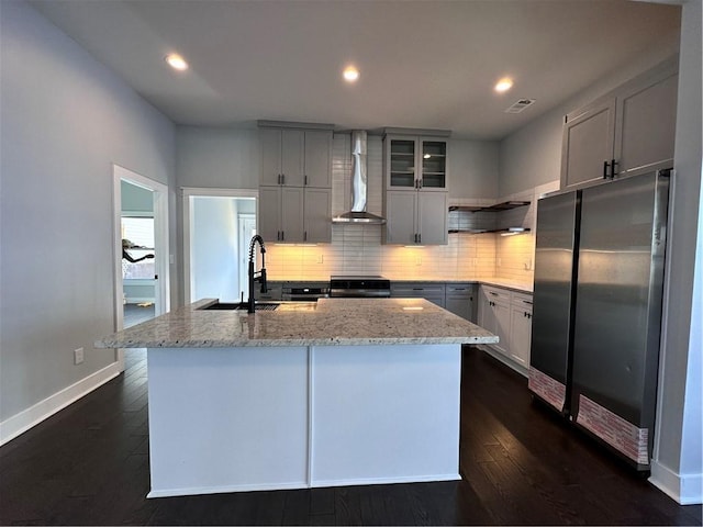 kitchen with wall chimney range hood, glass insert cabinets, a center island with sink, and freestanding refrigerator