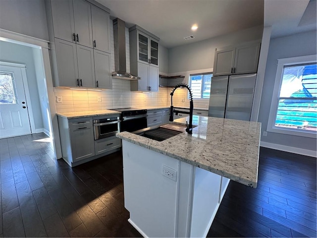 kitchen featuring glass insert cabinets, appliances with stainless steel finishes, decorative backsplash, wall chimney exhaust hood, and a center island with sink