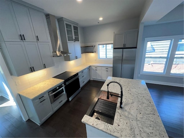 kitchen with light stone counters, backsplash, glass insert cabinets, appliances with stainless steel finishes, and wall chimney range hood