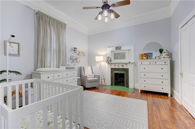 bedroom featuring a crib, a ceiling fan, wood finished floors, crown molding, and a fireplace