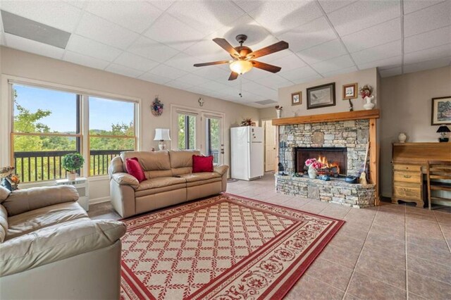 tiled living room with a ceiling fan, a stone fireplace, and a drop ceiling