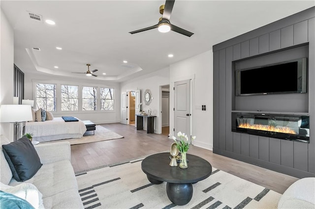 living area featuring a glass covered fireplace, a raised ceiling, light wood finished floors, and recessed lighting